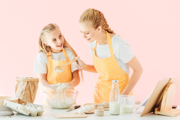 smiling young mother and adorable little daughter in yellow aprons kneading dough together isolated on pink - Foto, afbeelding