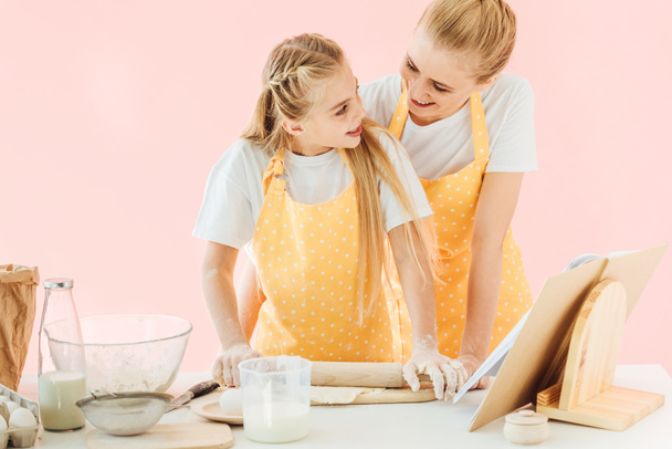 happy young mother and daughter kneading dough with rolling pin together isolated on pink - Photo, Image