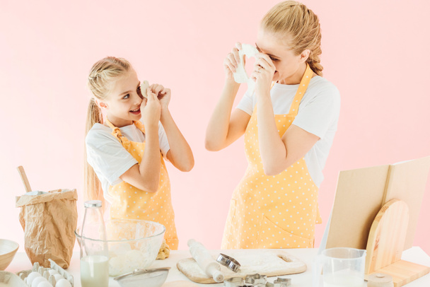 feliz madre e hija mirándose con trozos de masa mientras preparan galletas aisladas en rosa
 - Foto, Imagen