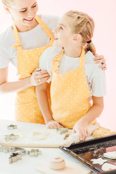 smiling mother and daughter preparing dough for cookies together and looking at each other isolated on pink - Photo, Image