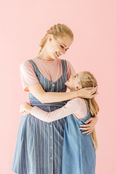 happy mother and daughter in blue dresses embracing and looking at each other isolated on pink - 写真・画像