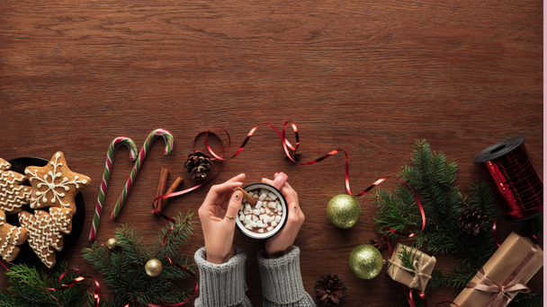 partial top view of person holding cup with hot chocolate and marshmallows, cookies, candy canes and christmas decorations on wooden background  - Fotó, kép