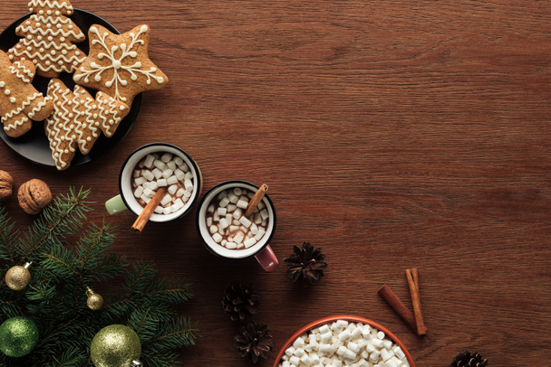 flat lay with cups with hot chocolate, marshmallows and cinnamon sticks, tasty cookies and fir twigs with shiny baubles on wooden surface, christmas background - Photo, Image