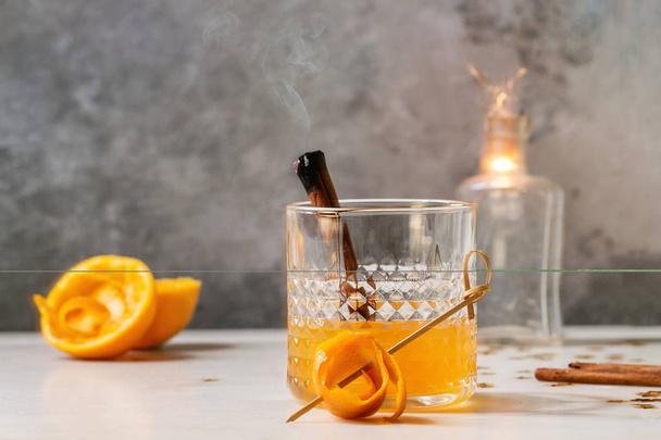 Glass of Scotch Whiskey orange juice alcohol cocktail with swirled orange peel and smoking cinnamon sticks standing on white marble table with golden holiday Christmas stars confetti and sparkler. - Foto, Bild