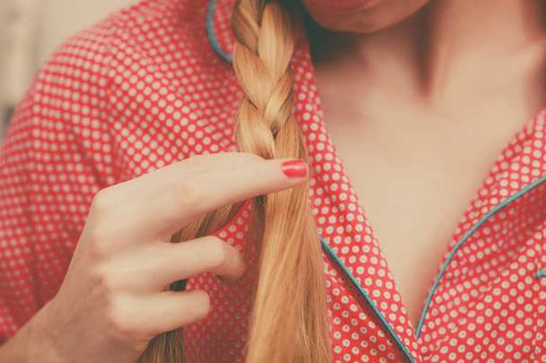 Fancy trendy blond hairstyle at home concept. Woman wearing pink pajamas doing braid on blonde hair - Fotó, kép
