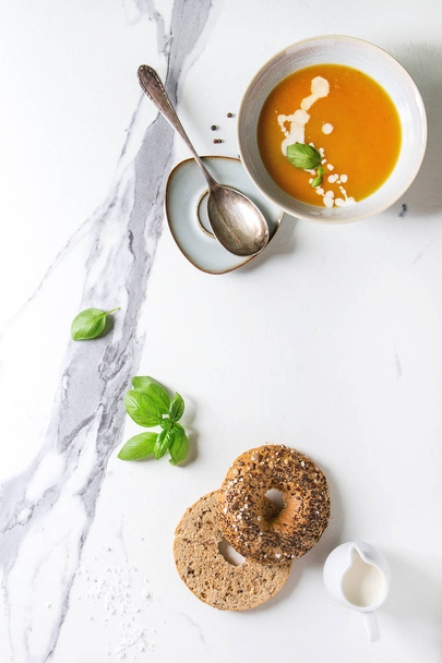 Bowl of vegetarian pumpkin carrot soup served with herbs, spoon, jug of cream, bagel bread, black pepper, salt over white marble texture background. Flat lay, space - Foto, Imagem