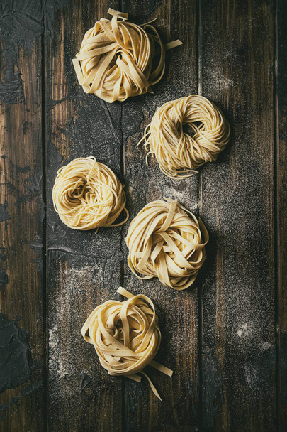 Variety of italian homemade raw uncooked pasta spaghetti and tagliatelle with semolina flour over dark plank texture wooden table. Flat lay, space - Фото, изображение
