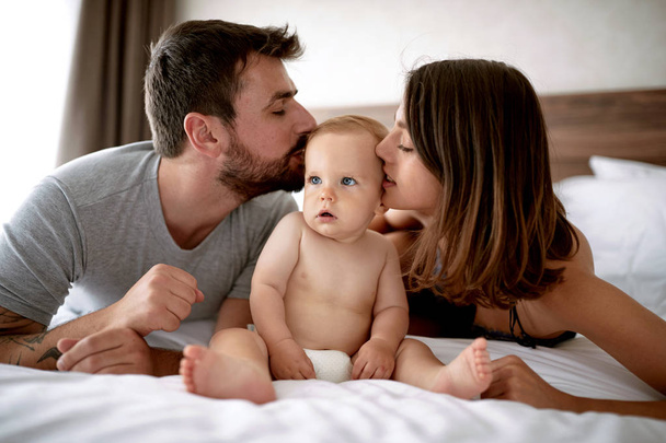 Семья с ребенком утром. Молодая мать, отец целует ребенка на белой кровати
 - Фото, изображение