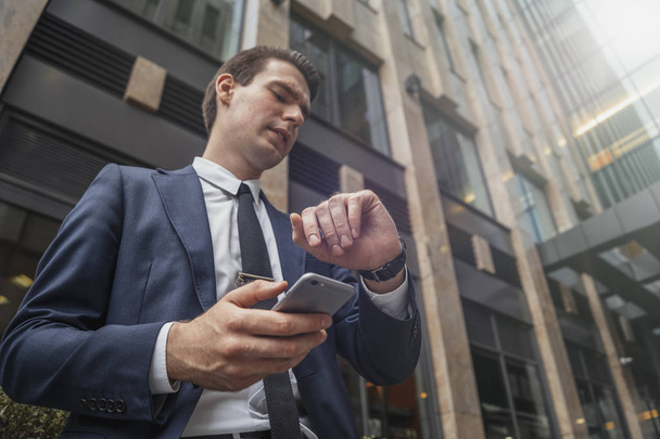 Κοντινό πλάνο του επιχειρηματία δίπλα στο επιχειρηματικό κέντρο στο κέντρο της πόλης κρατώντας το κινητό στο χέρι και βλέπουν ρολόγια. Κάτω όψη - Φωτογραφία, εικόνα