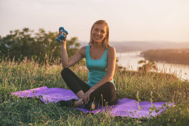 Beautiful sporty woman holding weights while sitting on exercise mat with a cityscape and river behinde her.Image is intentionally toned. - Foto, Bild