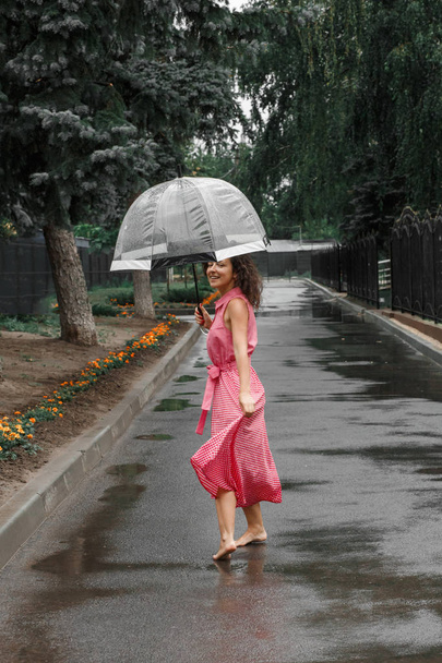 Jeune fille en robe rouge avec un parapluie transparent dansant sous la pluie debout dans une flaque d'eau
. - Photo, image