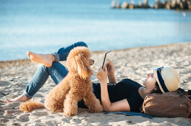 Woman traveler is holding touch pad, while is relaxing outdoors during his trip on the sea. Woman is traveling with her dog. - Foto, Imagem