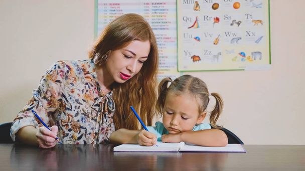 Little cute girl sits in classroom and studies with teacher in exercise book - Foto, Bild