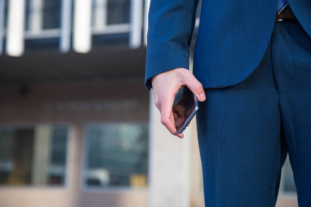 Gesichtslose Aufnahme eines Mannes im schicken Anzug, der sein Handy hält, während er mit der Hand in der Tasche auf der Straße steht. - Foto, Bild