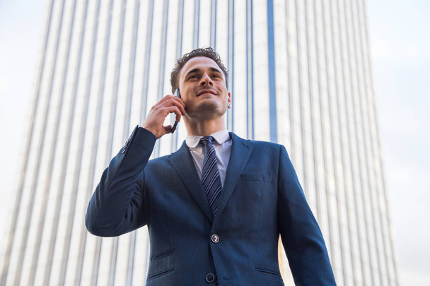 Веселый стильный бизнесмен в формальном костюме и с телефонными разговорами позирует на городском фоне
. - Фото, изображение
