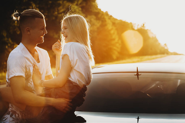 Vista lateral do casal alegre abraçando e inclinando-se no porta-malas do carro na beira da estrada em backlit
  - Foto, Imagem