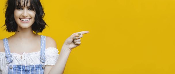 Lovely young woman smiling and looking at camera while pointing at empty space on bright yellow background - Foto, afbeelding