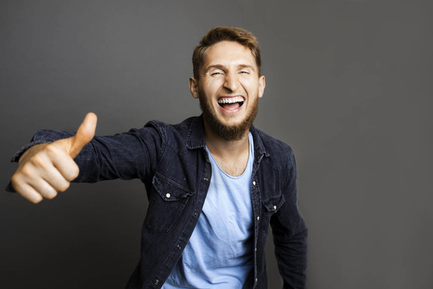 Handsome young guy laughing and showing thumb-up gesture while standing on gray background - Fotó, kép