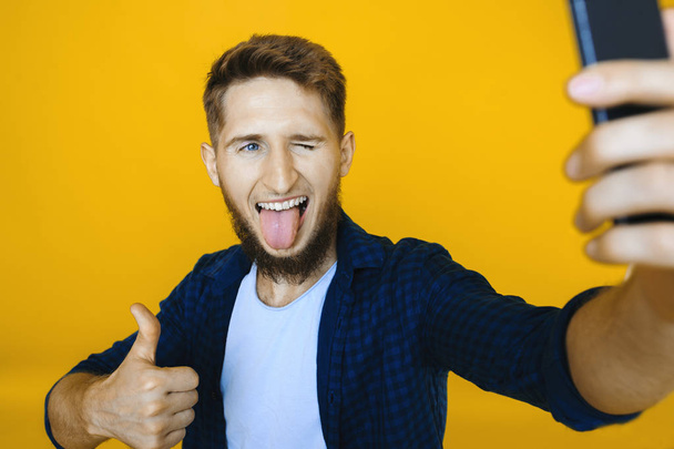 From above shot of young male showing tongue and gesturing thumb-up while posing for selfie on yellow background - 写真・画像
