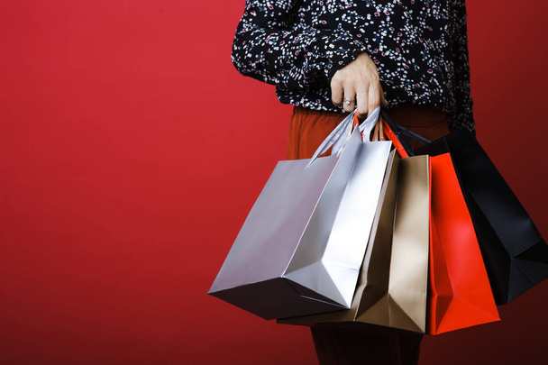 Crop view of slim attractive young woman in patterned shirt holding shopping bags of different color on red background - 写真・画像
