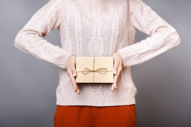Vue recadrée de la femme en pull tricoté chaud et pantalon en terre cuite tenant dans les mains présentes dans une petite boîte en carton sur fond gris
 - Photo, image