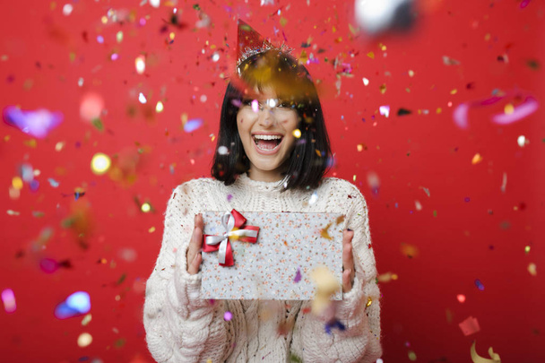 Young woman in knitted wool sweater standing in falling confetti and holding present on red background  - Фото, зображення