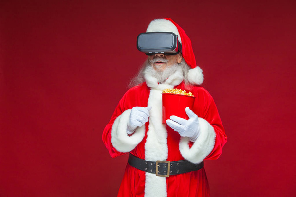 Christmas. Santa Claus in black virtual reality glasses holding a red bucket of popcorn. Watching a movie in virtual reality. Isolated on red background. - Zdjęcie, obraz
