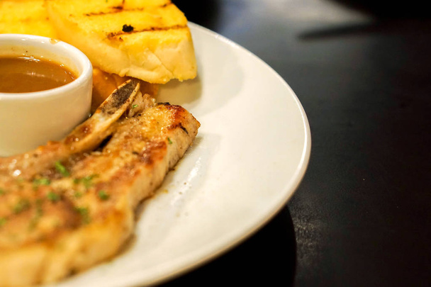 クローズ アップのレストランの黒いテーブルの白いセラミック プレートにペッパー ソース t-bone 豚肉ステーキとニンニク パンをトリミングし、. - 写真・画像