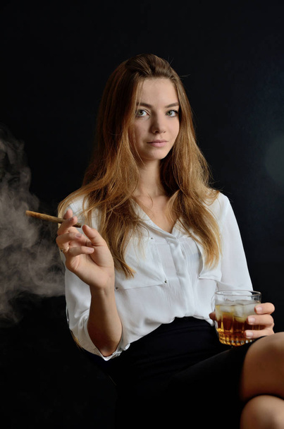 Young female sits with cigarillo and glass of whiskey in her hand. Elegant woman with white top and black dress. - Φωτογραφία, εικόνα