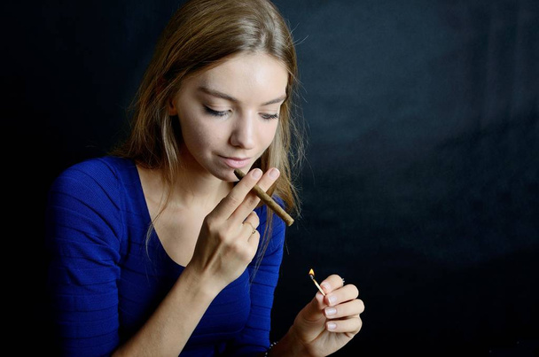 Young female lighting up cigarillo with matches. Woman in blue dress with black background. - Фото, зображення