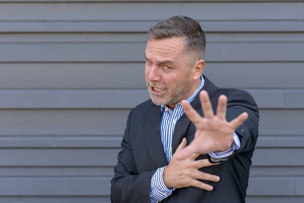 Бизнесмен делает жест остановки одной рукой, прикрывая грудь другой, как бы пытаясь оттолкнуть кого-то от серой стены с копировальным пространством
 - Фото, изображение