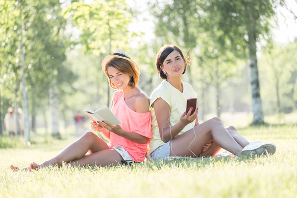 Two cheerful girls sitting on lawn, reading book, listening music on smartphone in green park at sunny warm day. Outdoor time spending in summer day - Foto, afbeelding