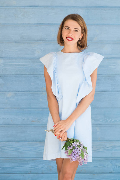 Beautiful portrait of young charming girl with bouquet of fresh lilac against blue wooden background - Foto, afbeelding