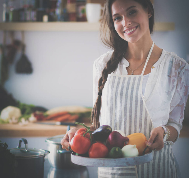 Smiling young woman holding vegetables standing in kitchen - Foto, imagen