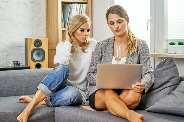 zwei junge und glückliche Frauen sitzen auf einer Couch mit einem Computer und surfen auf Webseiten - Foto, Bild