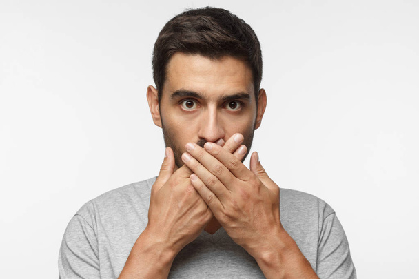 Secret, silence concept. Close up portrait of young man in gray t-shirt covering his mouth with hands isolated on grey background - Foto, Imagen