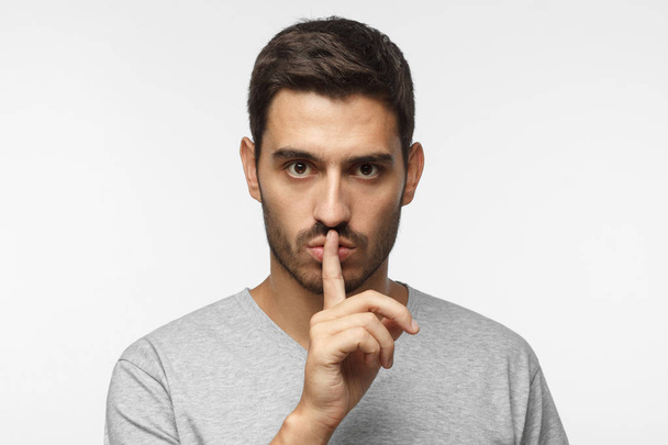Shhh gesture. Young man isolated on gray background dressed in white casual t-shirt pressing finger to lips as if asking to keep silenc - Foto, Imagen
