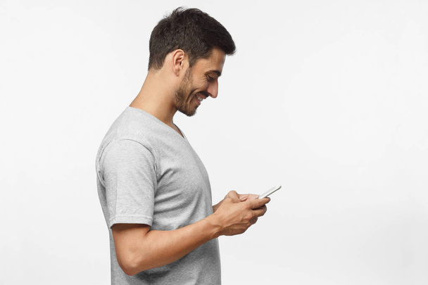 Side portrait of handsome man, isolated on gray background, looking down at smartphone screen, exchanging messages or surfing internet - Foto, Bild