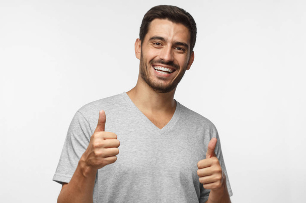 Young laughing man isolated on grey background showing thumb sup with positive emotions and happiness. Satisfaction with quality and recommendation - Foto, Imagem