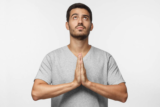 Younng man praying with hands pressed together, looking upwards as if asking god for blessing, isolated on gray background - Foto, imagen