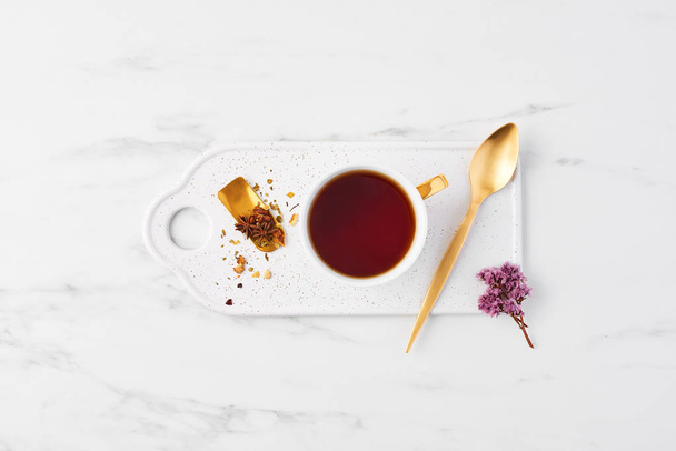Top view of white cup of tea with herbs on tea mesure and golden spoon on serving plate. Cup of tea on white marble background with dried flowers. Flat lay. - Photo, Image