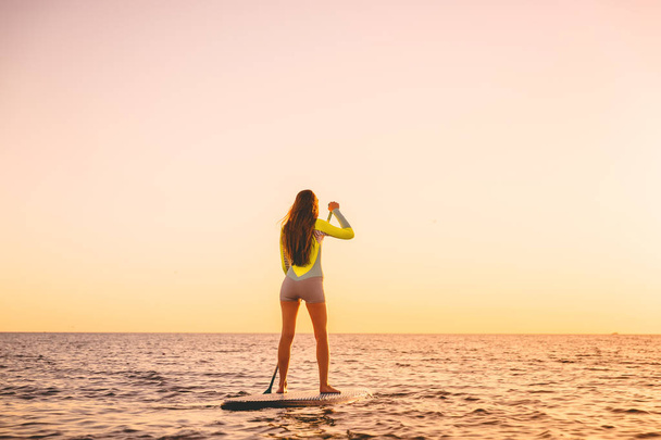 Genç kadın kürek kurulu sıcak günbatımı renkleri ile ayağa, yüzen - Fotoğraf, Görsel