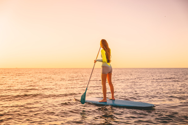 Jonge vrouw drijvend op stand up paddle board met warme kleuren van de zonsondergang - Foto, afbeelding