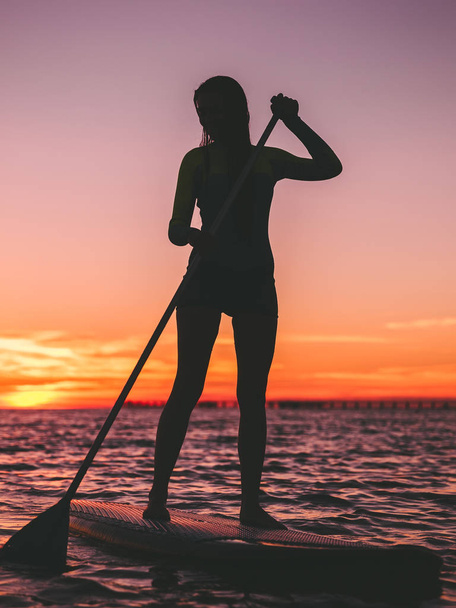Genç kadın kürek kurulu sıcak günbatımı renkleri ile ayağa, yüzen - Fotoğraf, Görsel