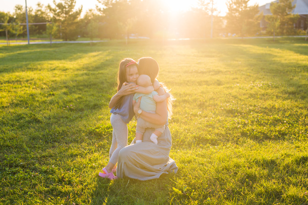 Mooie vrouw met dochter en baby zoon op groen grasveld. Moeder met twee kleine kinderen knuffelen buitenshuis. Gelukkige familie concept - Foto, afbeelding