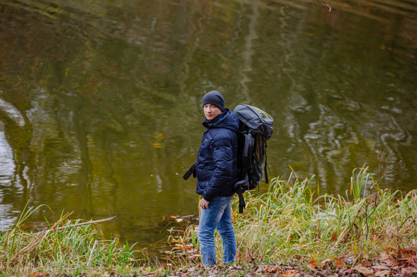 a tourist in a jacket and jeans looks at the camera near the river on the background of an autumn landscape - Фото, изображение