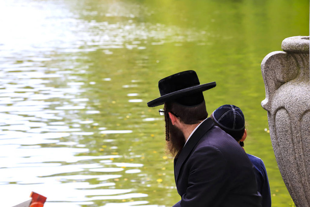 Сім'я хасидська євреїв, єврейський чоловік з хлопчиком, традиційні чорний одяг, сидячи на березі озера в парку, Умань, Україна - Фото, зображення