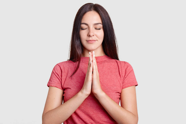 Pretty woman prays for good wellness, keeps palms pressed together, eyes shut, believes in good fortune, dresed in casual pink t shirt, poses against white background. People and faith concept - Photo, Image