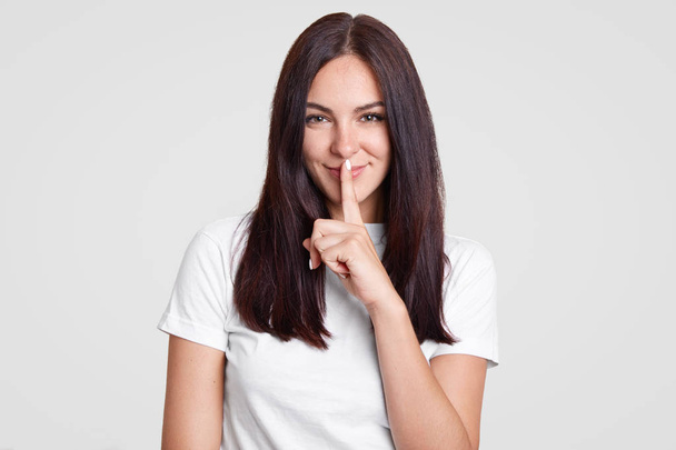 Attractive woman with long dark hair has secret, keeps fore finger over mouth, shows shush gesture, has satisfied facial expression, dressed in casual outfit, isolated over white background. - Photo, image