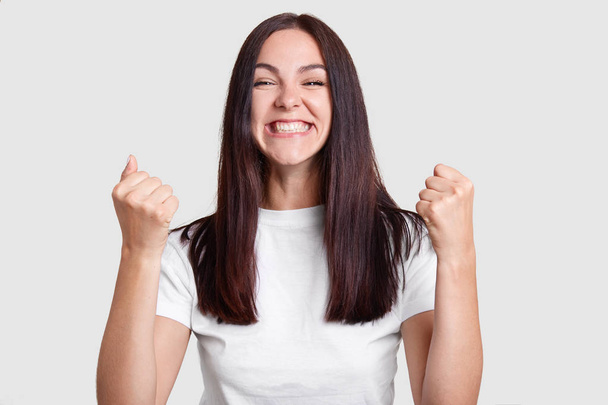 Successful woman clenches fists with happiness, has broad smile, squints face, dressed casual t shirt, isolated over white background. Carefree brunette girl rejoices success. Victory, determination - Photo, image